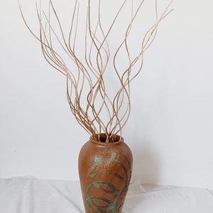 May include: A brown ceramic vase with a textured surface and green accents. The vase is filled with thin, brown, curly branches.
