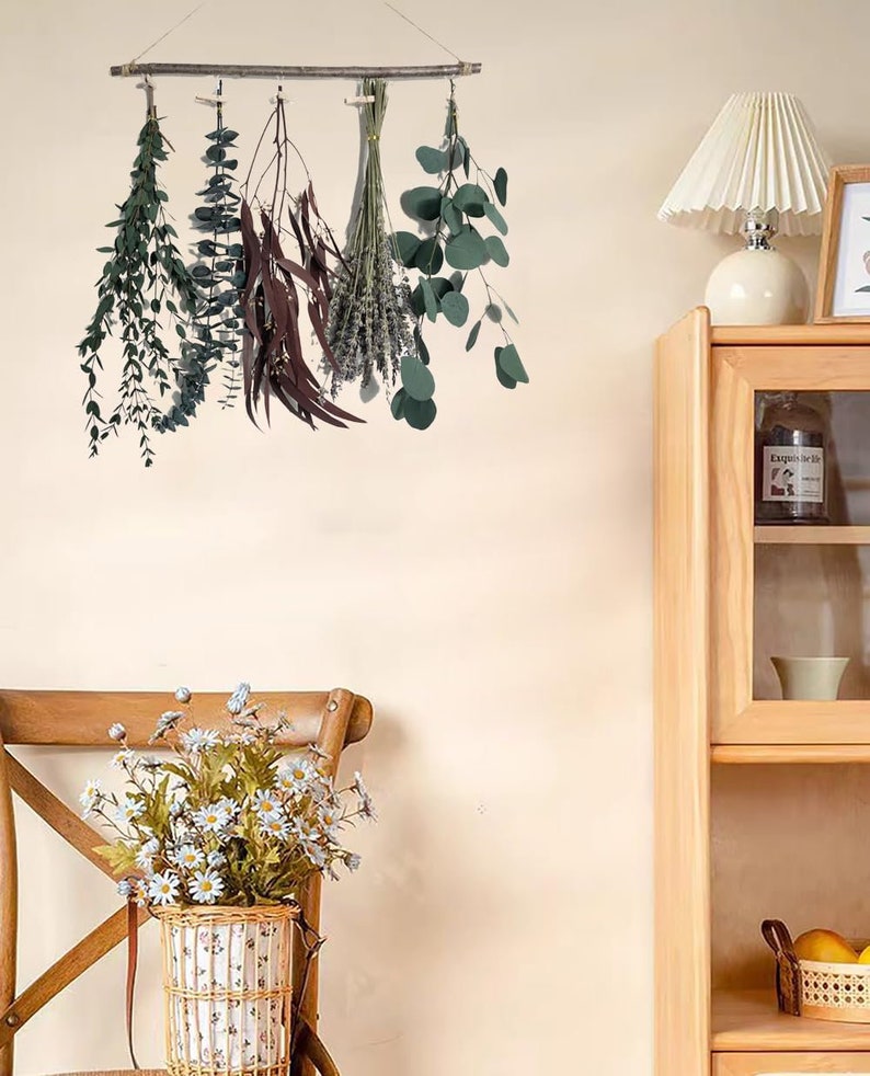 May include: A wooden dowel rod with five bundles of dried greenery hanging from it. The bundles include eucalyptus, lavender, and other leafy plants. The dowel rod is hanging on a white wall.