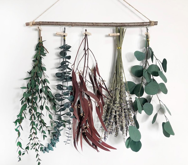 May include: A wooden dowel rod with five bundles of dried plants hanging from it. The bundles include eucalyptus, lavender, and red and green foliage.