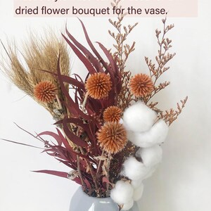 May include: A rustic-style dried flower bouquet in a grey vase. The bouquet includes dried wheat, cotton bolls, and burgundy eucalyptus leaves.