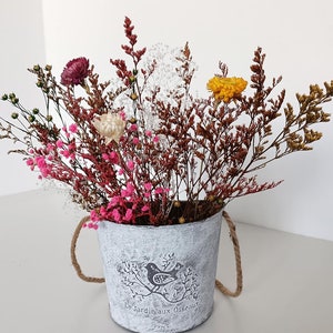 May include: Dried flower arrangement in a white metal bucket with the text "Le Jardin aux Oiseaux" on the side. The arrangement includes pink, white, and yellow flowers, and brown, green, and white stems.