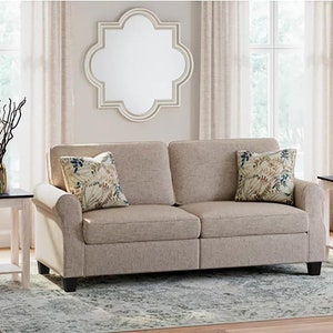 May include: A beige sofa with two floral patterned throw pillows sits in a living room with two white end tables with dark brown tops. A large, white, octagonal mirror hangs on the wall behind the sofa.