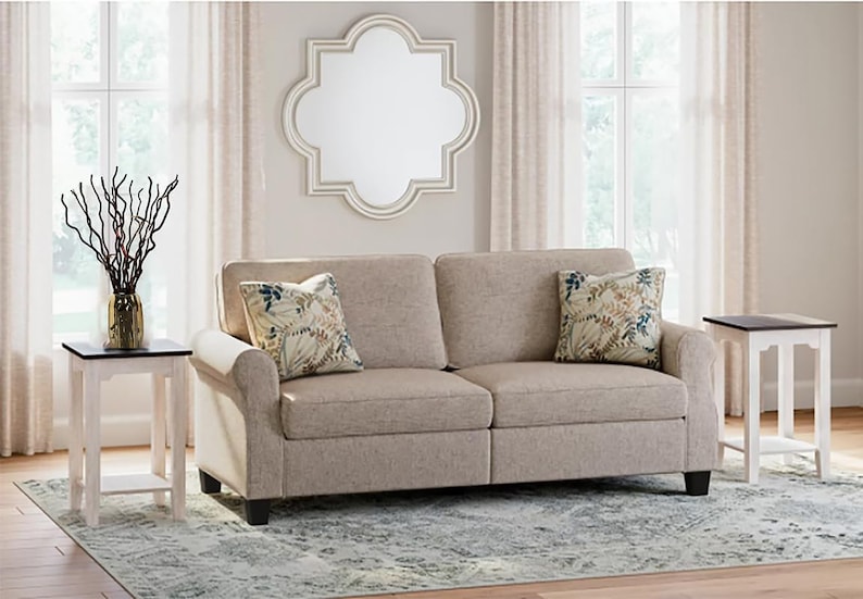 May include: A beige sofa with two floral patterned throw pillows sits in a living room with two white end tables with dark brown tops. A large, white, octagonal mirror hangs on the wall behind the sofa.