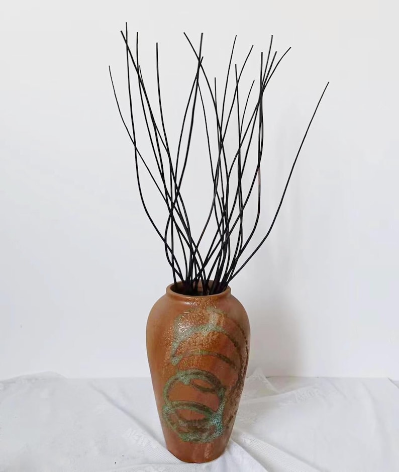 May include: A brown ceramic vase with a textured surface and green and brown swirling designs. The vase is filled with black twig-like branches.