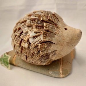 May include: A decorative hedgehog ornament with a textured, layered body in shades of brown and beige. The hedgehog has a smooth, light-coloured face and rests on a stone base with a small green leaf. A charming home decor item.