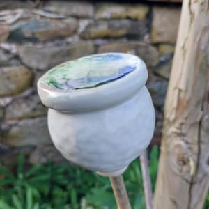 May include: A ceramic poppy seed head garden ornament. The white, textured sphere is topped with a flat, glazed disc with blue and green colours. It is mounted on a thin, brown stem. The background is blurred. The ornament is suitable for outdoor use.