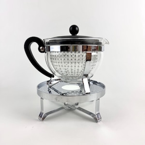 May include: A clear glass teapot with a black handle and lid, featuring a perforated metal infuser insert inside. The teapot rests on a circular chrome-plated metal stand with four legs and a perforated top surface for heat distribution.