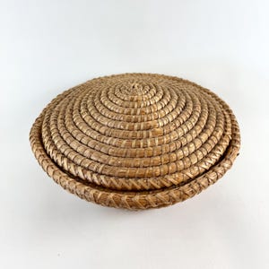 Puede incluir: Una cesta redonda tejida con tapa. La cesta est&aacute; hecha de paja natural y tiene un color marr&oacute;n claro.