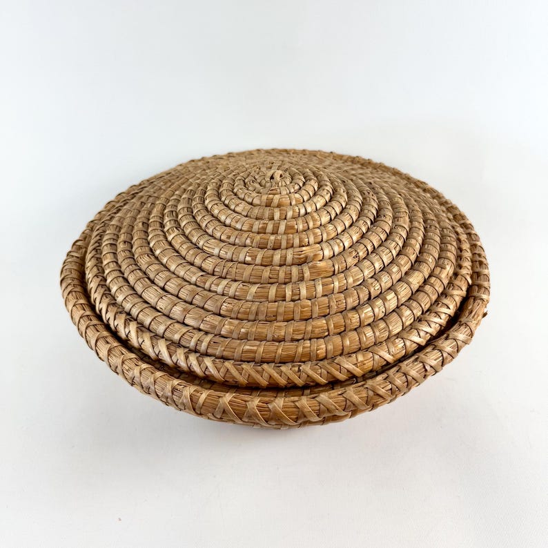 Puede incluir: Una cesta redonda tejida con tapa. La cesta est&aacute; hecha de paja natural y tiene un color marr&oacute;n claro.