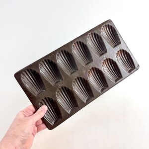 Op de afbeelding: Een rechthoekige, donkerbruine madeleine-vorm met 12 schelpvormige vormen. De vorm wordt door een hand tegen een witte achtergrond gehouden. De vormen hebben geribbelde details, perfect om te bakken.