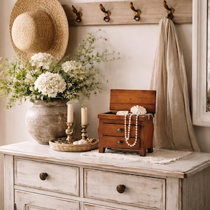 May include: A rustic wooden jewelry box with two drawers and an open lid, displaying a pearl necklace and a decorative flower. The box sits on a white dresser, alongside a vase of white flowers, candles, and a straw hat.