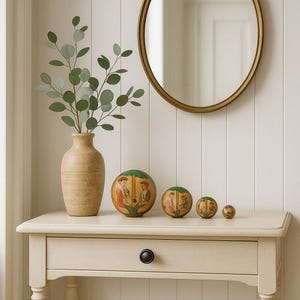 Puede incluir: Un jarrón beige con ramas verdes y frondosas se encuentra sobre una mesa color crema. Cuatro esferas decorativas de madera con figuras pintadas están dispuestas frente al jarrón. Un espejo ovalado con marco dorado cuelga de la pared blanca.