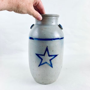 Op de afbeelding: Een grijze steengoed pot met een blauw sterontwerp. De pot heeft een brede opening en een rond lichaam.