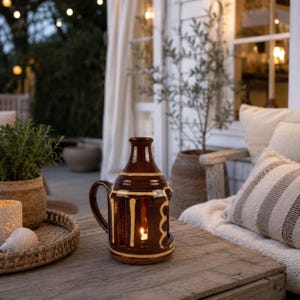 May include: A brown ceramic lantern with a glossy finish and vertical cream-coloured stripes. The lantern features a handle on one side and an open arched design that reveals a small, lit tea light candle inside, casting a warm glow from within.