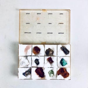 May include: An open, vintage mineral specimen box with a clear lid. The box contains sixteen individual compartments, each holding a different mineral sample. The lid is labeled with mineral names. The samples vary in color and texture, including quartz, pyrite, and malachite.