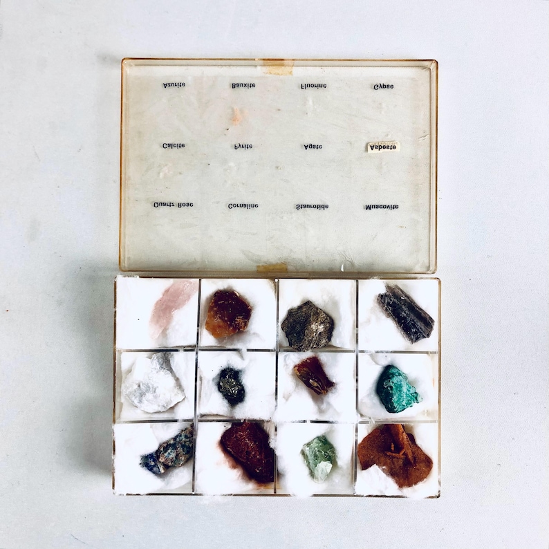 May include: An open, vintage mineral specimen box with a clear lid. The box contains sixteen individual compartments, each holding a different mineral sample. The lid is labeled with mineral names. The samples vary in color and texture, including quartz, pyrite, and malachite.