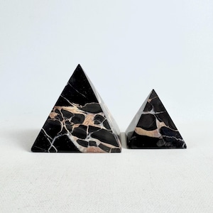 Könnte beinhalten: Zwei schwarze Marmorpyramiden mit beigen und weißen Adern. Die Pyramiden sind unterschiedlich groß, die größere links. Die Pyramiden sind dreieckig und haben eine polierte Oberfläche. Sie stehen auf einer weißen Oberfläche.