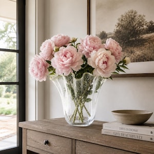 May include: A clear glass vase with a ruffled, wavy rim and a textured, bubbled surface containing a large bouquet of soft pink peony flowers. The vase sits on a wooden surface next to two stacked books with the text This is Home and Live Beautifully.