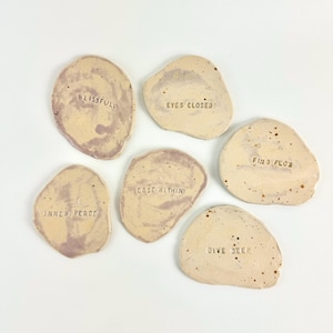May include: Six small, round, beige ceramic coasters with a light purple marbling effect. Each coaster has a different phrase embossed on the surface: "Blissful", "Eyes Closed", "Find Flow", "Inner Peace", "Ease Within", and "Dive Deep".