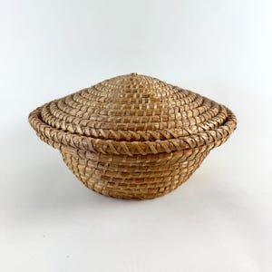 Puede incluir: Una peque&ntilde;a cesta redonda tejida con tapa. La cesta est&aacute; hecha de paja natural y tiene un color marr&oacute;n claro.