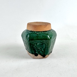 Puede incluir: Un pequeño tarro de cerámica verde con tapa. El tarro tiene forma hexagonal y está decorado con un patrón floral. La tapa está hecha de un material marrón.