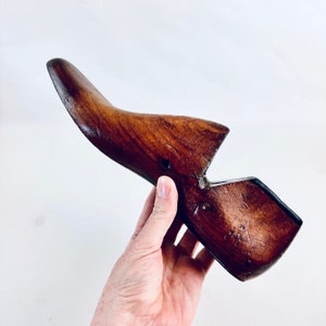 May include: A vintage wooden shoe last, with a curved toe and heel. The shoe last is a rich, dark brown colour, with visible wood grain and a smooth finish. The tool is held by a person's hand.