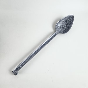 Könnte beinhalten: Ein langstieliger Löffel mit einer gesprenkelten grau-weißen Emaille-Oberfläche. Der Löffel hat eine tiefe, ovale Laffe und einen geraden, schlanken Stiel, der in einem leicht gebogenen Ende ausläuft und eine texturierte, gefleckte Optik aufweist.