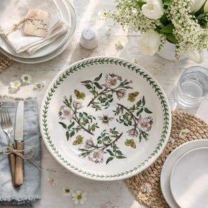 Può includere: Una ciotola da portata in ceramica bianca con un design floreale e di insetti. La ciotola presenta un bordo di foglie verdi e un motivo centrale di fiori rosa, farfalle gialle e un'ape. La scena è su un tavolo di legno rustico.