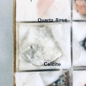 May include: A display case with a white calcite mineral sample. The sample is labeled "Calcite" in black text. The case also contains a "Quartz Rose" sample and other mineral specimens.