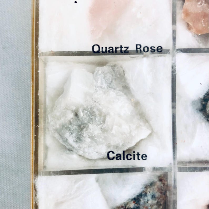 May include: A display case with a white calcite mineral sample. The sample is labeled "Calcite" in black text. The case also contains a "Quartz Rose" sample and other mineral specimens.