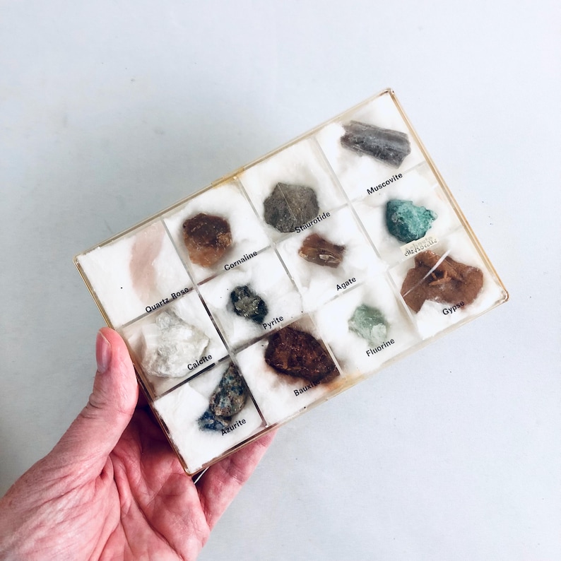 May include: A collection of various minerals and gemstones displayed in a clear plastic case. The case contains specimens labeled Quartz Rose, Calcite, Azurite, Pyrite, and others. The minerals are in a variety of colors, including brown, green, and white.