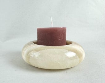 May include: A white alabaster candle holder with a round, slightly oval shape. A small, dark pink candle sits inside the holder.