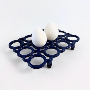 以下が含まれることがあります： 丸いデザインの濃い青色の金属製エッグホルダーで、2つの白い卵が入っています。ホルダーには小さな足と格子状のパターンがあります。卵は楕円形で、表面にテクスチャがあります。背景は白です。