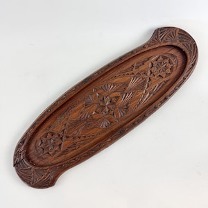 May include: A long, oval-shaped wooden tray with intricate carved designs. The tray is made of dark wood and features a raised edge.