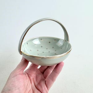 Könnte beinhalten: Ein kleiner, hellgrüner Keramikkorb mit Henkel, Goldrand und goldenen Sternmustern. Der Korb wird in einer Hand gehalten und unterstreicht sein zartes Design und seine Vintage-Ästhetik.