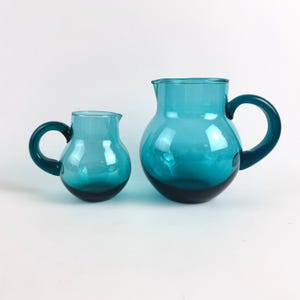 Op de afbeelding: Twee blauwgroene glazen kannen van verschillende groottes, elk met een rond lichaam, een brede schenktuit en een gebogen handvat. Het glas is doorschijnend met een gladde, glanzende afwerking en vertoont een consistente diepblauw-groene kleur.