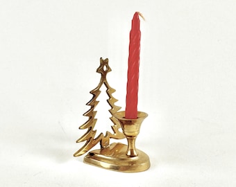 May include: A polished brass candle holder featuring a cutout silhouette of a Christmas tree with a star at the top. The base supports a single, slender, twisted red taper candle that stands upright in the centre of the metal holder.