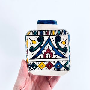 Puede incluir: Un jarrón de cerámica blanca con forma cuadrada y un patrón geométrico azul, verde, amarillo y rojo. El jarrón está decorado con diseños florales estilizados.