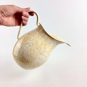 May include: Cream-coloured pitcher with a speckled pattern of white and yellow. The pitcher has a curved handle and a flared spout. The pitcher is held by a hand against a white background. The pitcher is likely used for serving beverages.