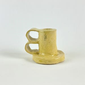 Peut inclure: Un petit bougeoir en céramique jaune fait à la main. Le bougeoir a une base ronde, un corps cylindrique et une poignée à deux boucles. La pièce est émaillée et a un aspect rustique et artisanal.