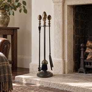 Pode incluir: Um conjunto de ferramentas para lareira com um suporte central de base circular que sustenta um atiçador, uma pá e uma escova. As ferramentas possuem hastes de metal escuro e cabos de latão envelhecido em formato de pinhas decorativas.
