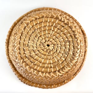 Puede incluir: Una cesta redonda tejida de paja natural de color marr&oacute;n claro. La cesta tiene una forma circular y est&aacute; tejida en un patr&oacute;n apretado e intrincado.
