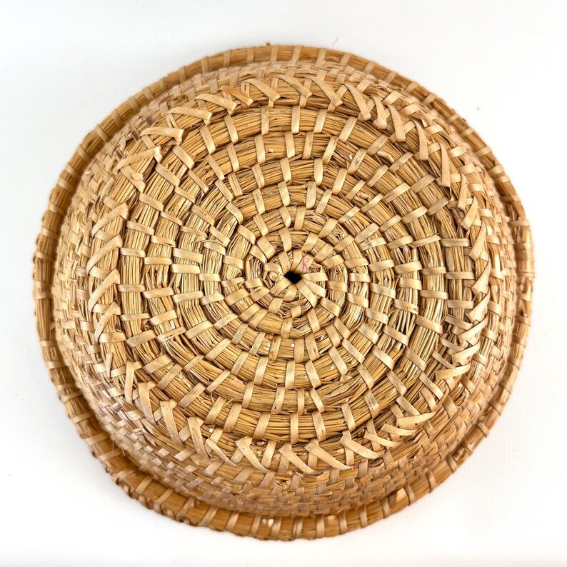 Puede incluir: Una cesta redonda tejida de paja natural de color marr&oacute;n claro. La cesta tiene una forma circular y est&aacute; tejida en un patr&oacute;n apretado e intrincado.