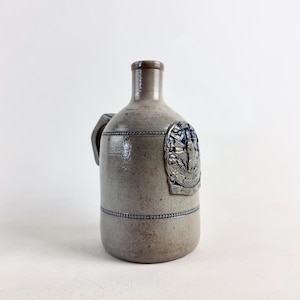 May include: A stoneware jug with a handle and a decorative blue and silver emblem. The jug is a light gray color with a brown neck and a dark blue band around the body. The emblem features a detailed design.
