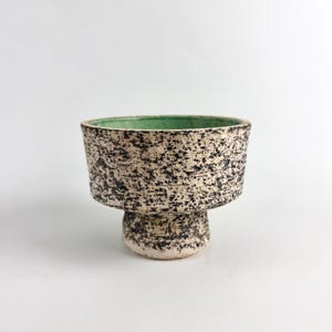 May include: A set of three ceramic mugs with a speckled, matte finish. The mugs are in shades of cream, light gray, and dark gray. Each mug has a simple, minimalist design and a rounded handle.