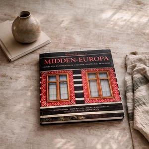 May include: A book titled "MIDDEN-EUROPA" with a red and black cover design featuring two window illustrations. A small, round, beige vase sits atop a stack of books. A linen cloth is on the right.