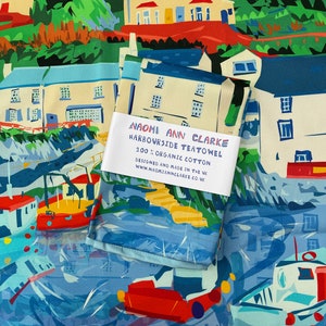 Può includere: Un asciugamano da tè colorato con una scena di città costiera. L'asciugamano è realizzato in cotone biologico al 100% e presenta il testo "NAOMI ANN CLARKE HARBOURSIDE TEATOWEL 100% ORGANIC COTTON DESIGNED AND MADE IN THE UK WWW.NAOMIANNCLARKE.CO.VK".