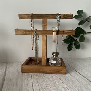 May include: Wooden jewellery stand with two tiers for necklaces and bracelets. The stand also has a small tray for rings or other small items. A watch and a bottle of perfume are displayed on the stand.