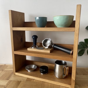 May include: A wooden shelf unit with three shelves, holding various coffee brewing tools and accessories. The top shelf has two bowls, one green and one blue. The middle shelf has a tamper, a portafilter, and a wooden base. The bottom shelf has a funnel, a tamper, and a stainless steel jug.