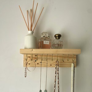 May include: A wooden jewellery organiser with brass hooks, displaying necklaces and earrings. A white ceramic diffuser with reeds and two perfume bottles are on the shelf above. The jewellery includes a gold chain, a turquoise floral necklace, and a gold chevron necklace.